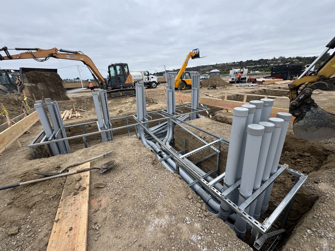 Conduit stub-ups racked on strut frames in open trenches while an excavator backfills